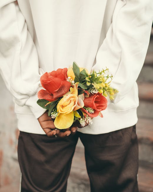 Person in a white hoodie is holding a vibrant bouquet of fresh flowers behind their back. The arrangement includes large, coral and yellow roses, along with smaller yellow and white blooms, complement