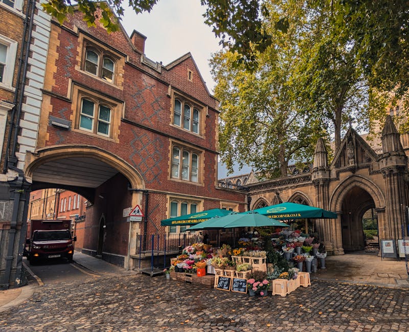 A vibrant outdoor flower stall situated on a cobbled street in front of a historic brick building with an arched gateway and Gothic architectural details. The stall is shaded by a large green umbrella