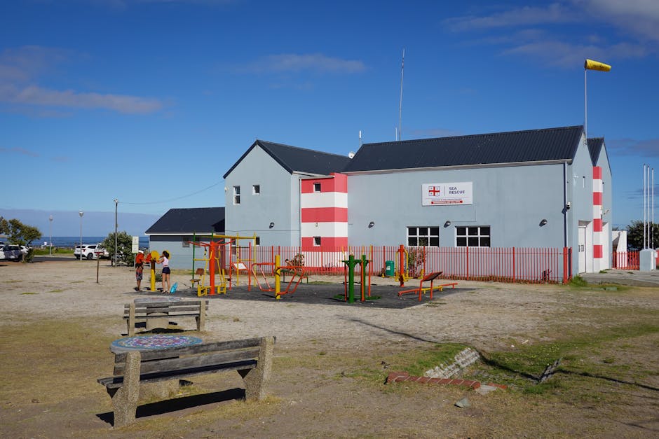 The image features a large, light gray building with red and white striped accents, labeled as a sea rescue center, situated outdoors under a bright blue sky with scattered clouds. In the foreground,