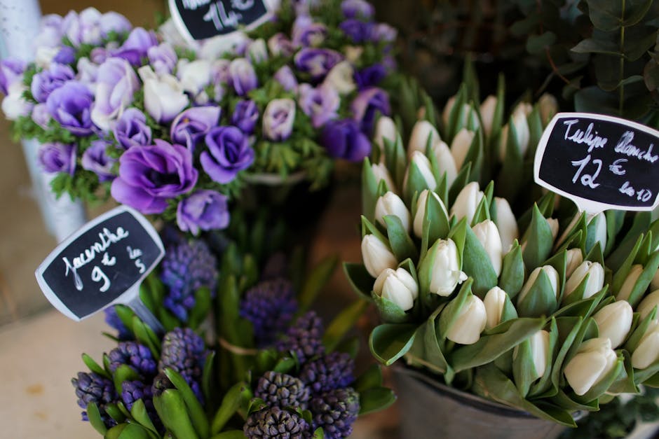 A vibrant display of fresh flowers arranged in glass and ceramic containers at a florist shop. The arrangement features white tulips with closed petals, surrounded by green leaves, set against a backg
