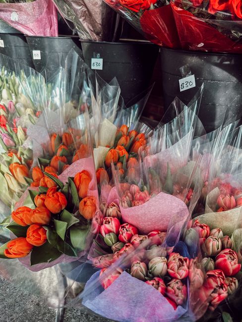 A display of various flower bouquets arranged in black buckets at a florist shop. The bouquets feature vibrant tulips in shades of orange, pink, and red, wrapped in clear and pink tissue paper, with s