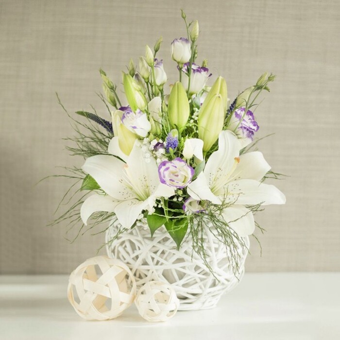 The photograph focuses on an elegant floral arrangement that would make a refined wedding table centerpiece in Motspur Park, combining soft colour with thoughtful structure. At the base is a rounded white vase whose surface is formed from an intricate open-weave pattern, reminiscent of finely woven wicker or rattan, creating a feeling of lightness and modern craft. From this bowl-like container, stems of white Asiatic and oriental-style lilies stretch and curve, some flowers already fully open with flared, trumpet-shaped petals and central stamens carefully arranged, others still held tight in slender green buds that rise above the arrangement with a sense of anticipation. Nestled among the lilies are blooms of Eustoma (lisianthus) in white and cream, their delicate petals tipped or veined with touches of lavender-violet, which introduce a gentle, romantic tone suited to a wedding breakfast near Motspur Park's quiet residential streets. Deeper purple or blue accent flowers are tucked between these main blooms, offering a cool note and visual depth. The bouquet is enriched with an abundance of greenery: feathery, almost cloud-like fronds mingle with more substantial, glossy green leaves that overflow slightly at the edges of the vase, giving a relaxed, garden-gathered feel. Tiny white filler florets, much like baby's breath, punctuate the spaces between, adding softness and a sense of air in the composition. The vase stands on a clean, bright white surface, beside which sit two decorative woven spheres in shades of light beige or natural wood, echoing the container's lattice texture and extending the organic theme. Behind it, a softly lit, light beige wall with a subtle horizontal, linen-like texture provides an understated, warm backdrop that allows every flower and leaf to be clearly defined. Overall, the scene feels tranquil and celebratory, perfect for weddings, vow renewals, or meaningful gatherings in the Motspur Park area.
