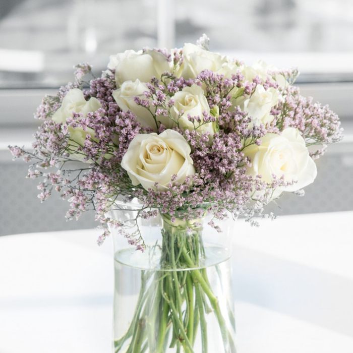 In this image, a sophisticated sympathy bouquet of white roses and lavender filler flowers takes centre stage, resting in a tall, clear glass vase that allows every detail of the stems and water to be seen. The roses are plentiful and fully open, their petals smooth and velvety, unfolding in controlled spirals that create rounded, substantial blooms. Their colour is a serene blend of bright white and soft ivory, suggesting purity, remembrance, and quiet reflection. Encircling and threading between these focal blooms are abundant sprigs of tiny lavender and soft pink flowers, probably limonium or statice, whose minute, star-shaped blossoms cluster together to form airy, cloud-like masses. These pastel accents introduce gentle colour and texture, softening the edges of the arrangement while preserving its understated elegance. Through the transparent glass vase, the long green stems are neatly aligned and submerged in clear water, where they intersect in delicate, organic lines that echo the bouquet's refined composition. The vase rests on a white or very pale surface that reflects some of the natural light, casting faint highlights that enhance the arrangement's sense of freshness. Behind it, the background fades into a smooth blur of whites and light greys, reminiscent of a calm interior near Motspur Park's tree-lined streets and small gardens. The overall mood is still and contemplative, as though the bouquet has been placed gently beside a framed photograph, a condolence card, or a memorial candle. This is the sort of arrangement a local Motspur Park family might choose to honour a loved one, its gentle tones and careful balance offering solace and a quiet reminder of enduring love.