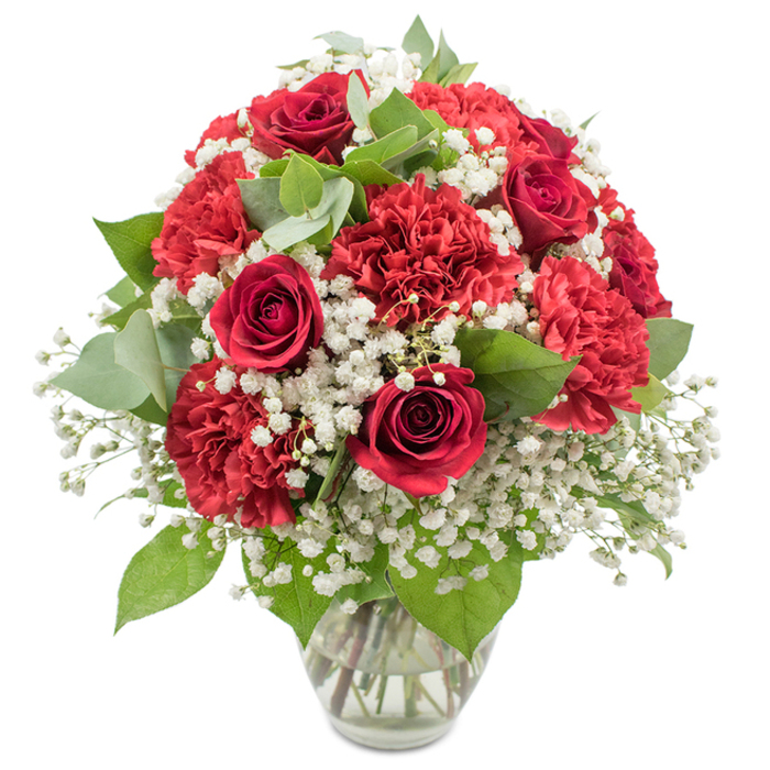 A classic red and white floral arrangement is captured in a clear glass vase, positioned against a bright white backdrop that isolates the bouquet and brings its colours into sharp relief. Several deep, velvety red roses form the focal point, each bloom tightly furled into an elegant spiral that suggests both restraint and intensity, much like the quiet yet close-knit community around Motspur Park. Encircling these roses is a generous mass of red carnations; the petals are finely ruffled and layered, creating a plush surface that gives the bouquet a full, rounded, dome-like silhouette. Interspersed among the rich reds are delicate stems of white baby's breath, their tiny blossoms clustering together to form soft, cloud-like patches that add light, airiness, and a sense of purity, gently breaking up the intensity of the crimson tones. Fresh greenery winds through the composition: dark, glossy rose foliage provides a sturdy base, while round, silvery-green eucalyptus leaves add subtle movement and a cool contrast against the warmer reds. The vase is straightforward and modern, with a slightly flared top and crystal-clear sides that reveal the neatly gathered stems resting in clear water, a testament to careful conditioning and expert arranging. The directional studio lighting is bright but soft, highlighting the textures of the rose petals and carnation frills without casting harsh shadows, making the bouquet feel both polished and welcoming. This arrangement's timeless red-and-white palette lends itself beautifully to anniversaries, Valentine's Day, and romantic surprises delivered across Motspur Park, from homes near West Barnes Lane to cosy flats tucked away on quiet side streets, where it can brighten a room and convey heartfelt emotion.