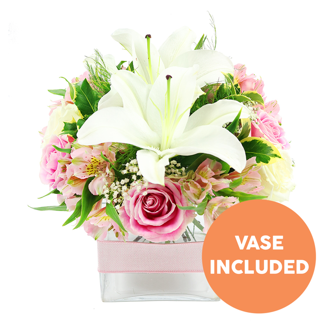 A stylish floral arrangement of white and pink flowers is captured in this image, standing out vividly against a bright, uncluttered white background. Dominating the centre are two large white Oriental lilies, fully open so their broad petals stretch wide and curl at the tips, revealing prominent reddish-brown stamens and a soft green core. These lilies create a striking focal point that contrasts beautifully with the surrounding softer tones. Around them, numerous pink roses gather in a compact cluster, some with tightly wrapped petals that hint at newly formed buds, others fully unfurled into lush, layered rosettes, adding a sense of romance and luxury. Threaded among the roses and lilies are stems of pink and white alstroemeria, their petals slender and slightly flared, many adorned with fine speckles and gentle yellow hints near the centre, bringing delicate pattern and texture to the piece. Tiny white blossoms of baby's breath drift lightly throughout, filling spaces between the larger blooms and giving the arrangement a misty, cloud-like softness. The entire bouquet is framed by varied greenery: smooth, glossy leaves offer a polished structure, while softer, feathery foliage introduces a subtle wildness, reminiscent of the informal gardens around Motspur Park and nearby New Malden. All stems sit securely in a low, square clear glass vase, whose transparency showcases the neatly arranged stems and clean water, conveying freshness and professional care. Around the middle of the vase, a pale pink satin ribbon is tied in a straight, horizontal band, adding a gentle decorative edge that makes the arrangement feel like a ready-made gift for a Motspur Park home or office. In the bottom right corner, a bright orange circular sticker with bold white text clearly states "VASE INCLUDED," signalling that the glass container is included with the flowers and ready to place directly on a table or windowsill.